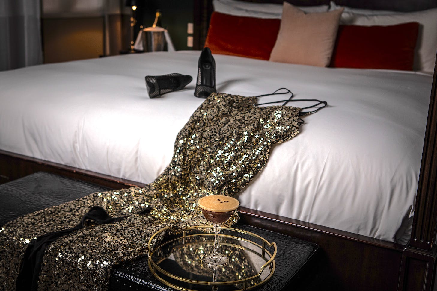 A sequinned dress rests on a hotel bed behind an espresso martini